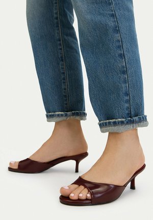 Stopy w ciemnoczerwonych mule na niskim obcasie typu kitten heel z odkrytymi palcami, zestawione z podwiniętymi niebieskimi jeansami na jednolitym tle.