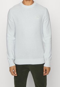 Hellblauer Strickpullover mit Rundhalsausschnitt, gerippten Bündchen und Saum, mit einem kleinen quadratischen Logo auf der Vorderseite. Weiche Textur und figurbetonter Schnitt.