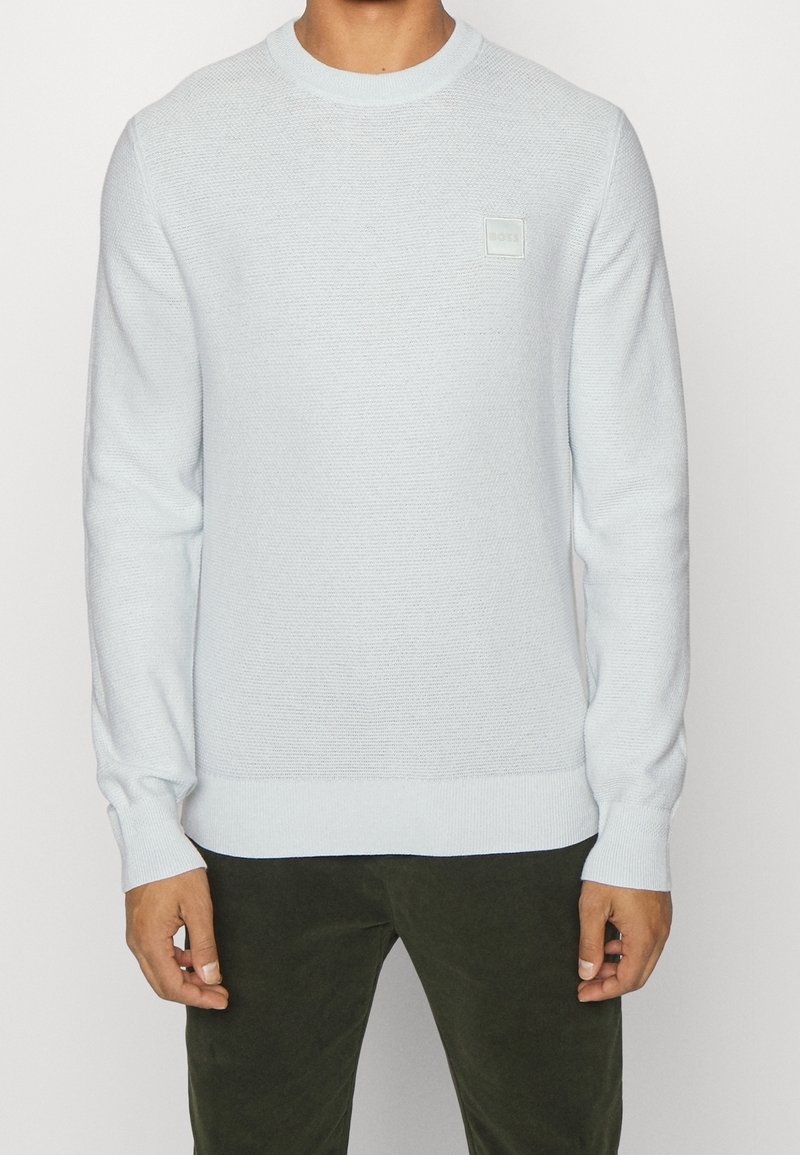 Hellblauer Strickpullover mit Rundhalsausschnitt, gerippten Bündchen und Saum, mit einem kleinen quadratischen Logo auf der Vorderseite. Weiche Textur und figurbetonter Schnitt.