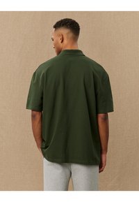 Tamsiai žalia trumpomis rankovėmis polo marškinėliai su klasikiniu apykakle ir laisvu siluetu, pagaminti iš lygaus audinio, rodomi iš nugaros.