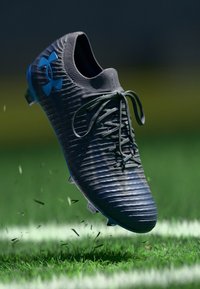 Scarpa da calcio nera con tomaia testurizzata, logo blu, lacci e suola trasparente, sollevata sopra un prato verde con detriti di erba sparsi.