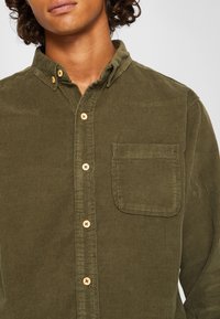 Camisa de pana verde oliva con cuello abotonado, que incluye un bolsillo en el pecho y botones de madera a lo largo de la tapeta frontal.