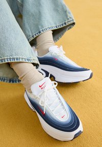 Le sneaker Nike Air Max 97 presentano una tomaia a sfumature blu e bianco, dettagli rossi e una suola spessa ammortizzata. Abbinate a calzini beige a coste.