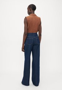 Chemise sans manches marron associée à un jean ample taille haute bleu foncé. Le tissu a une texture lisse avec des coutures visibles sur les poches.