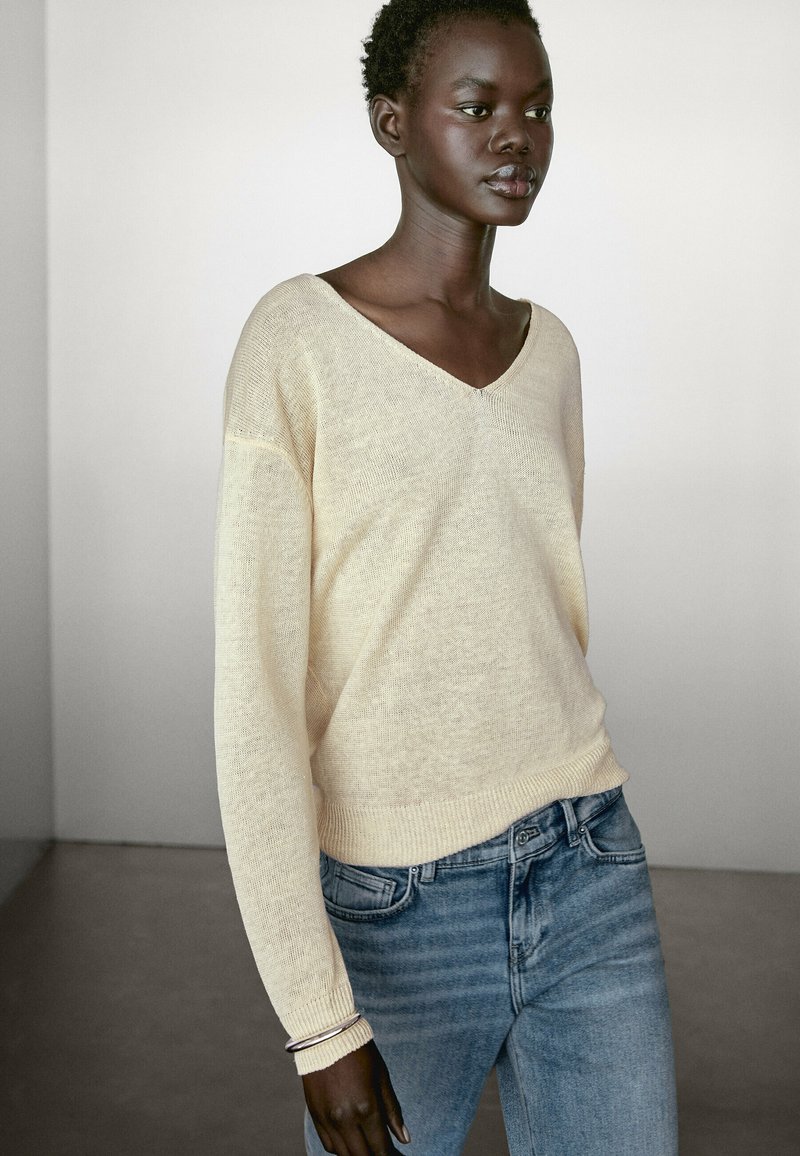 Maglione beige morbido a V con un fit ampio, maniche lunghe e polsini a costine, abbinato a jeans in denim azzurro chiaro con sottili righe.
