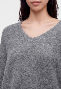 Femme portant un pull gris doux et texturé à col en V, montrant le décolleté et les épaules sur un fond neutre.