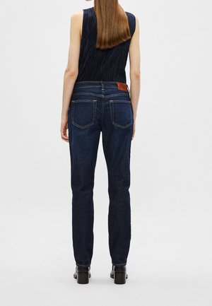 Femme aux longs cheveux raides portant un haut sans manches bleu foncé et un jean bleu foncé, debout de dos sur un fond uni.