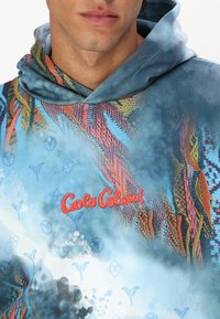 Hooded sweatshirt with vibrant multicolored abstract patterns. Made from soft fabric, features a logo in red and a blue color scheme.