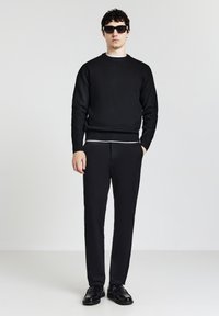 Schwarzer Strickpullover mit geripptem Saum über einem weißen Hemd, kombiniert mit schwarzen Hosen und schwarzen Schuhen. Der Träger trägt außerdem eine dunkle Sonnenbrille.