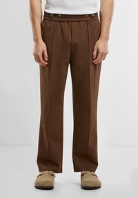 Brown, pleated pants with an elastic waistband and snap detailing. Fabric is smooth and lightweight. Paired with beige slip-on shoes.