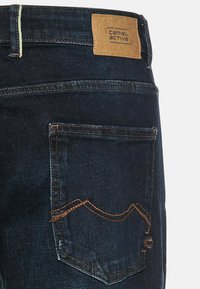 Donkerblauwe spijkerbroek van denim met een tan leren label, contrasterende stiksels en een uniek zakontwerp met gebogen stiksels.