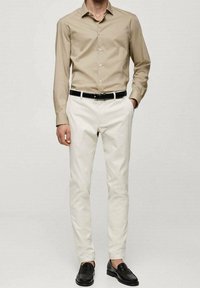 Camicia beige a maniche lunghe, pantaloni crema, cintura in pelle nera e mocassini neri. Texture lisce, design aderente e stile classico.