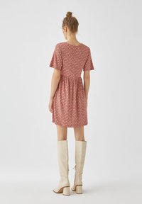 Rotes Blumenkleid mit kurzen Ärmeln und tailliertem Schnitt, aus leichtem Stoff. Knielang; kombiniert mit beige kniehohen Stiefeln.