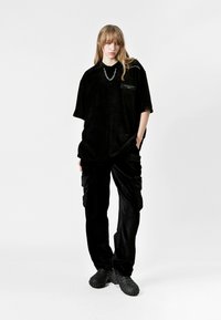 Top de micropana negro de gran tamaño con un bolsillo en el pecho y detalle de cadena, combinado con pantalones cargo de micropana negra y calzado texturizado negro.