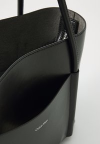 Borsa a mano in pelle nera con superficie liscia, design minimalista, apertura superiore e tasca interna visibile con logo del marchio.