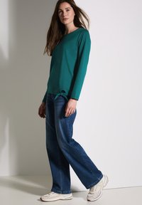 Teal blouse met lange mouwen en een zijstrikdetail, gecombineerd met blauwe wijde jeans en lichte sneakers, tegen een neutrale achtergrond.