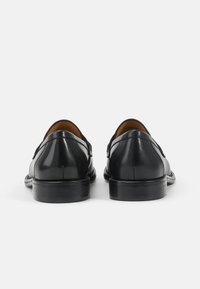 Chaussures en cuir noir avec une finition brillante, à talon bas et bout rond. Détails de couture et accents sur les côtés le long des coutures.