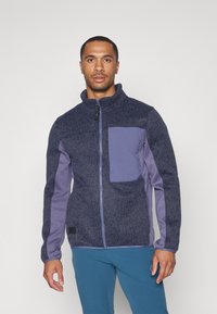 Veste en polaire bleu marine avec un corps en maille texturée et des manches violettes unies, dotée d'une fermeture éclair et de détails sur la poche avant.