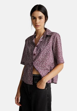 Femme portant une chemise violette à fleurs à manches courtes et boutons, avec un pantalon noir, une main dans la poche, sur un fond clair uni.