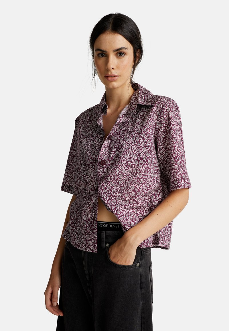 Donna che indossa una camicia floreale viola con maniche corte e bottoni, pantaloni neri, con una mano in tasca, su uno sfondo semplice e chiaro.