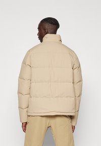 Beige gewatteerde jas met een hoge kraag, gepresenteerd vanuit de achterkant. Heeft een gestikte ontwerp en textuurstof voor isolatie.