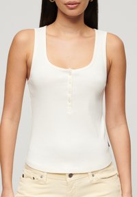 Hvid ribbet tanktop med en rund halsudskæring, der har en knaplukning og en tætsiddende pasform. Blød tekstur, velegnet til lag-på-lag.
