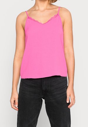 Kobieta w jasnoróżowej koronkowej bluzce na ramiączkach i czarnych, wysokich dżinsach, stojąca na jednolitym tle.