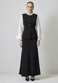 Černý elegantní vest s zlatými knoflíky přes bílé tričko, doplněný dlouhou černou sukní. Má stažený pas s šňůrkou. Černé vysoké podpatky.