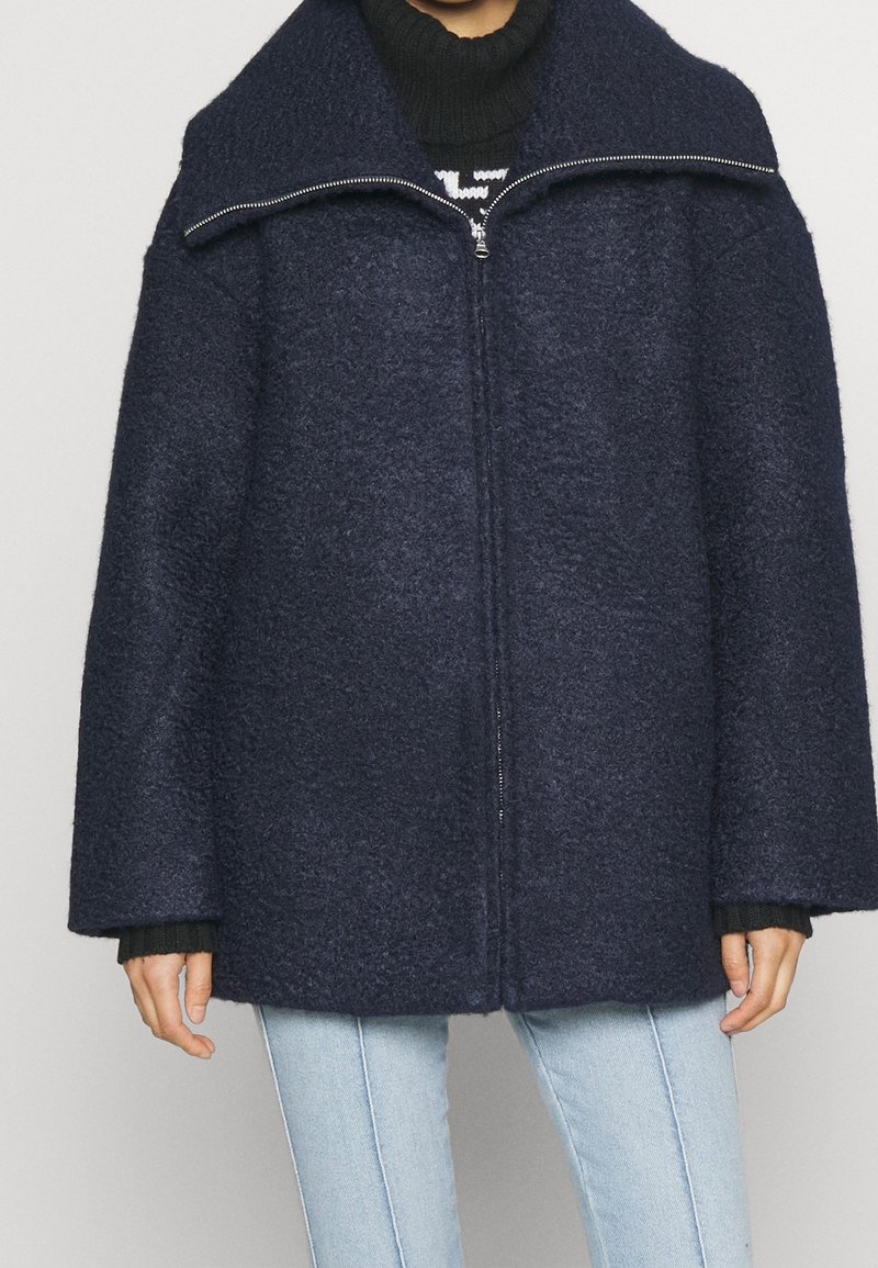 Marinblå ullblandning zip-up kappa med hög krage, oversized passform och texturert tyg, bärs över en svart ribbad tröja och ljusa jeans.