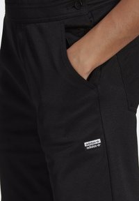 Černé látkové kalhoty s viditelnou boční kapsou a malým obdélníkovým bílým logem Adidas poblíž kapsy. Hladký povrch.