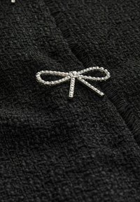 Čierna textúrovaná tkanina so striebornou brošňou v tvare mašle, zvýraznenou jasnými rhinestones, čo pridáva dekoratívny prvok k materiálu.