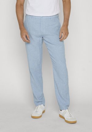 Mann trägt hellblaue Hose und weiße Sneaker, steht vor einem einfarbigen hellen Hintergrund.