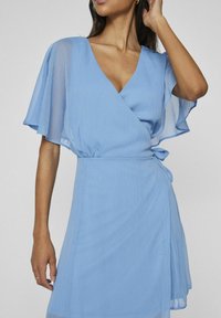 Svietlo modré zavinovacie šaty z textúrovanej látky. Majú krátke, voľne splývajúce rukávy, hlboký V-krk a opasok na páse.