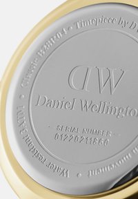 Apaļš pulksteņu aizmugure ar pulētu sudraba virsmu, uz kuras ir iegravēts teksts. Zelta krāsas metāla aplis ieskauj malu. Redzams sērijas numurs.