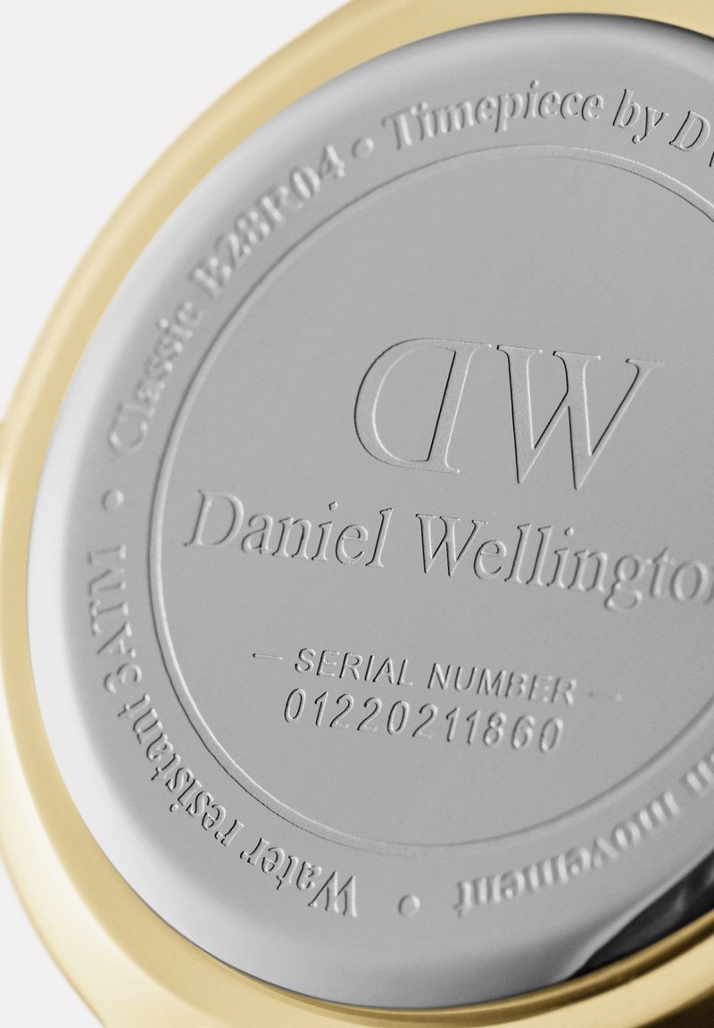 Runder Uhrenboden mit einer polierten Silberoberfläche und eingraviertem Text. Ein goldfarbenes Metallreifen umrahmt den Rand. Seriennummer sichtbar.