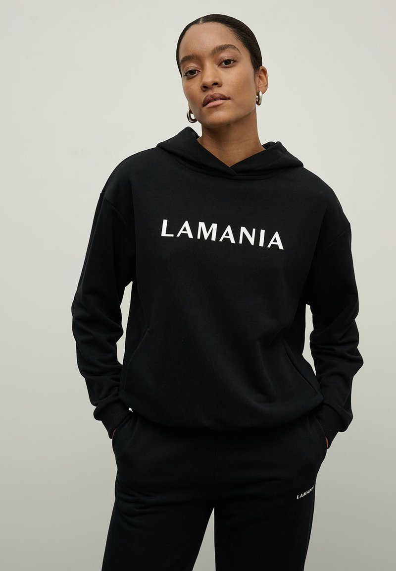 Juoda gobtuvinė džemperis su užrašu "LAMANIA" baltomis raidėmis. Minkšta medžiaga, laisvas siluetas ir rupūs rankogaliai. Derinamas su atitinkančiomis juodomis kelnių.