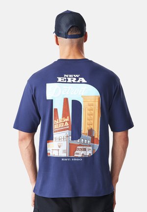 Hombre con camiseta y gorra New Era azul marino, vista trasera, la camiseta muestra un gráfico del skyline de Detroit con el texto "New Era Detroit" y "Est. 1920".