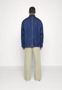 Denimjacka i djupblå färg, med en slät textur, krage och synlig sömnad. Matcher med vida beige byxor.