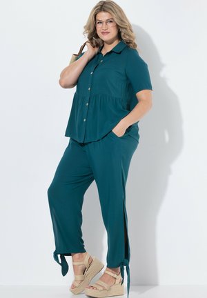 Kobieta o falujących blond włosach, ubrana w zieloną bluzkę na guziki i spodnie z wiązaniami przy kostkach, beżowe sandały na koturnie oraz okulary, stojąca na tle białego tła.