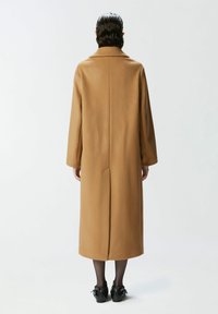 Dug košuljasti kaput u boji kamila s visokim ovratnikom, ravnim siluetom, bočnim džepovima i prorezom straga radi lakšeg kretanja.