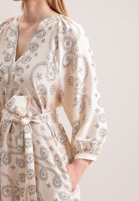 Krämfarbenes Kleid aus glattem Stoff, mit schwarzen Paisleymustern, tiefem V-Ausschnitt und gerafften Ärmeln mit einer gebundenen Taille.