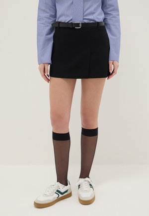 Femme portant une mini-jupe noire avec une ceinture, des chaussettes noires transparentes montantes, des baskets blanches et une chemise à manches longues rayée bleue rentrée.