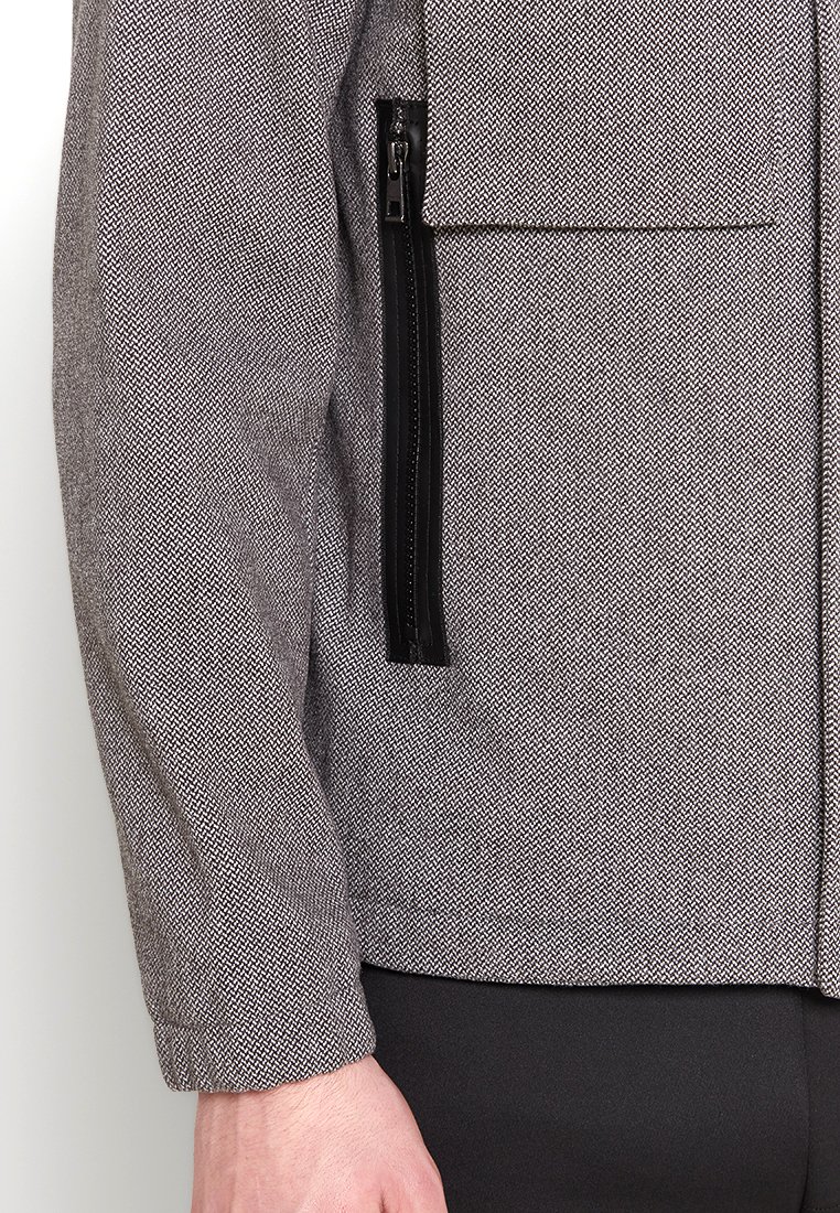 Grijze jas met een structuur en een visgraatpatroon, voorzien van zwarte ritsaccenten en een borstzak aan de linkerkant.