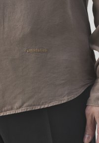 Camisa marrón tejida con una textura sutil, dobladillo por debajo de la cintura y un logo de la marca bordado en marrón oscuro en el lado izquierdo.