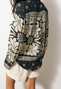 Koszula z długim rękawem w czarno-beżowy kwiatowy wzór, wykonana z lekkiego materiału, z zaokrąglonym dołem i kołnierzykiem zapinanym na guziki, w zestawie z beżowymi szortami.