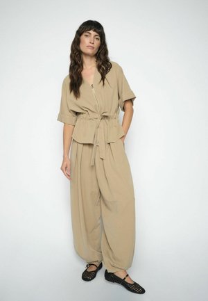 Woman with long wavy hair wearing loose beige zip-up top with tie waist and matching wide pants, paired with black woven shoes, standing against white.