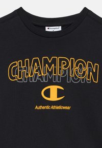 Μαύρο βαμβακερό T-shirt με έντονη κίτρινη επιγραφή "CHAMPION" και λογότυπο, με τη φράση "Authentic Athleticwear" σε μικρότερο μέγεθος κάτω από αυτήν.