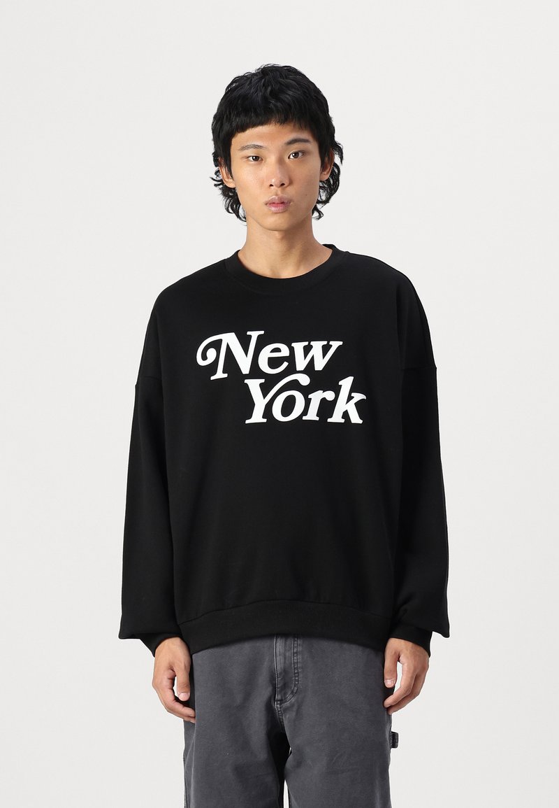 Čierna mikina s nápisom "New York" potlačeným bielou farbou, spájajúca okrúhly výstrih a dlhé rukávy. Hladká textura, uvoľnený strih.