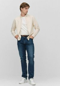 Licht beige overhemd met knopen over een wit T-shirt, gecombineerd met donkerblauwe spijkerbroek en witte sneakers met groene accenten.