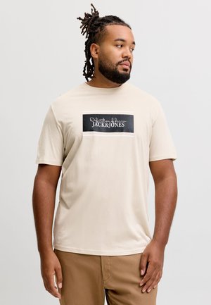 Mężczyzna z dredami, ubrany w beżowy t-shirt Jack & Jones i brązowe spodnie, stoi i patrzy w prawo na jednolitym tle.